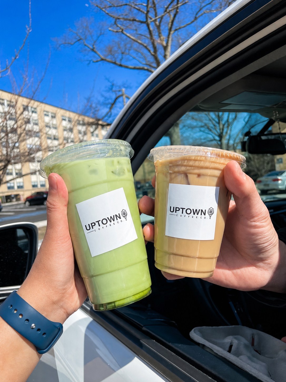 Two hands clinking an iced matcha and an iced latte with Uptown Coffee Co. labels at a car window