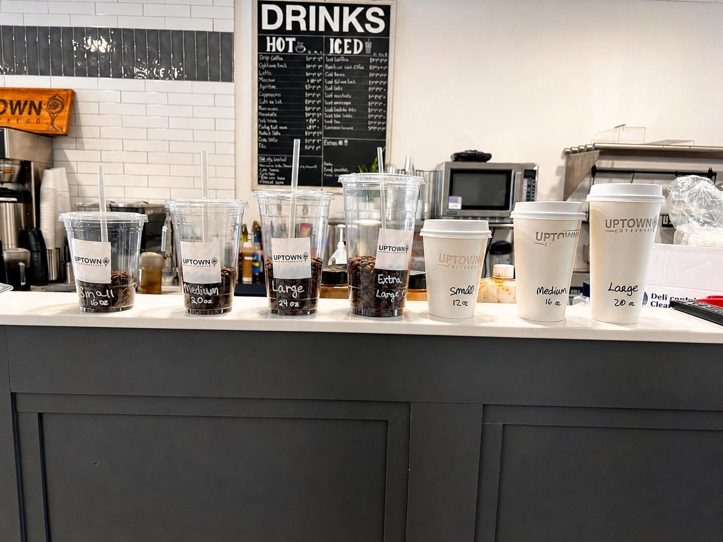 Uptown Coffee Co. hot and iced cup sizes lined up on the counter under the drinks menu board